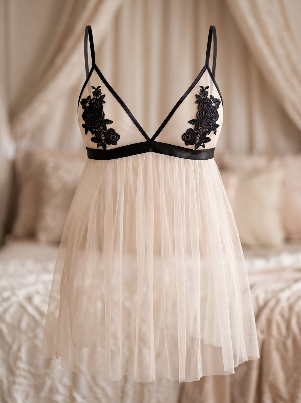 La nuisette blanche vue de face sur mannequin invisible, ses deux bonnets de voile transparent dont les galons noirs se croisent en X au milieu du buste, un bouquet de roses en dentelle sur chacun, et sa jupe évasée jusqu’à mi-cuisse