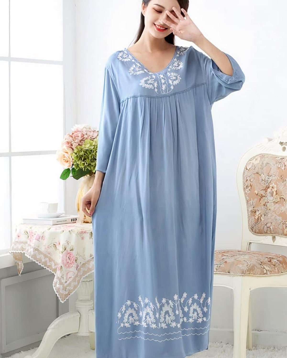 Nuisette longue bleue en coton brodé portée debout de face, main levée près du visage, broderies florales blanches sur le col en V