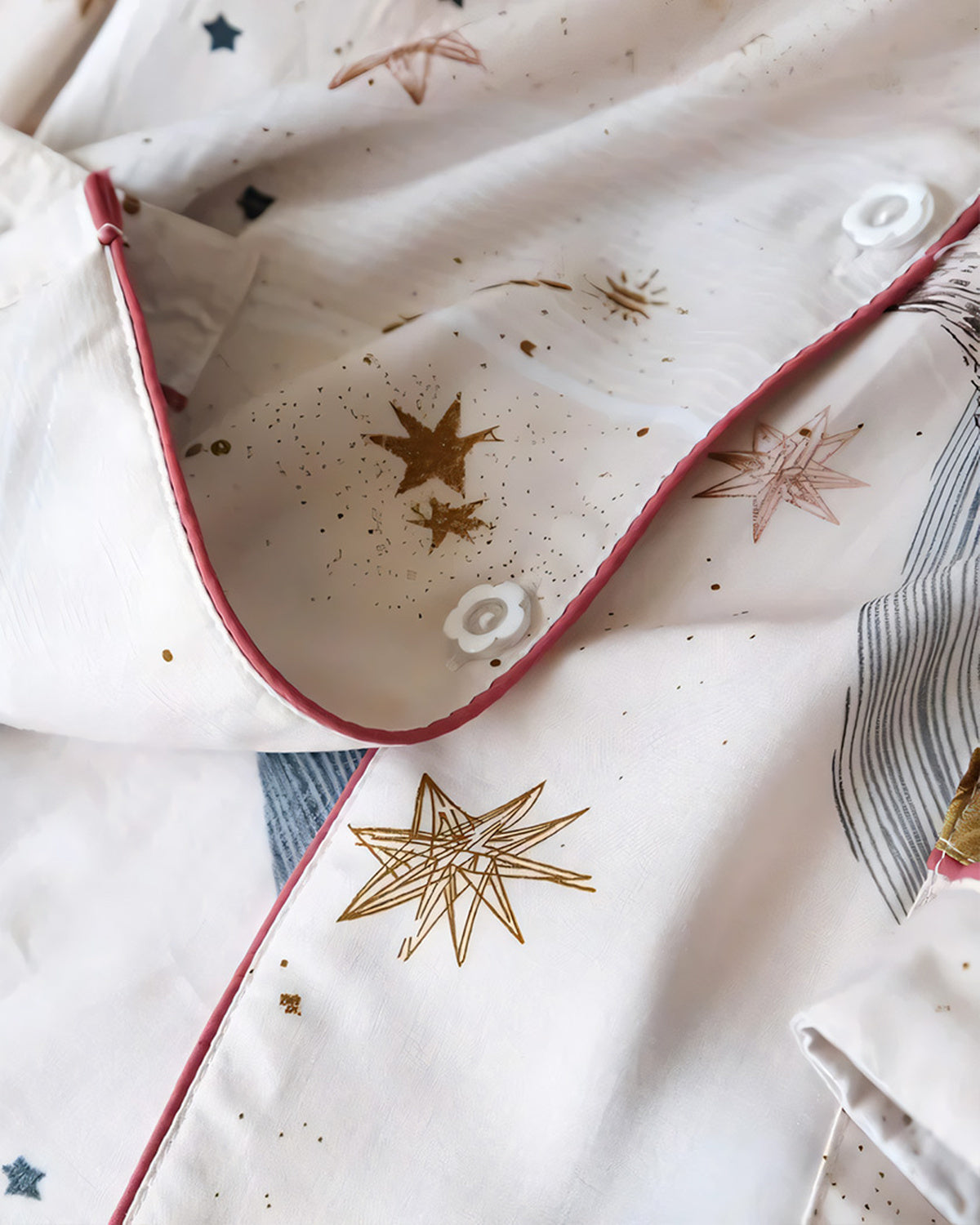 Gros plan sur le col arrondi d'une chemise de pyjama crème, son passepoil framboise, un bouton du devant et les étoiles dorées de l'imprimé