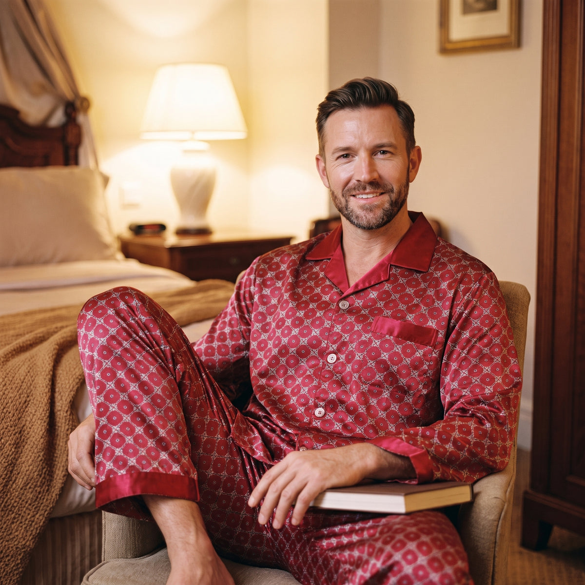 Homme installé dans un fauteuil le soir en pyjama satiné rouge, un livre ouvert sur les genoux et la lampe de chevet allumée