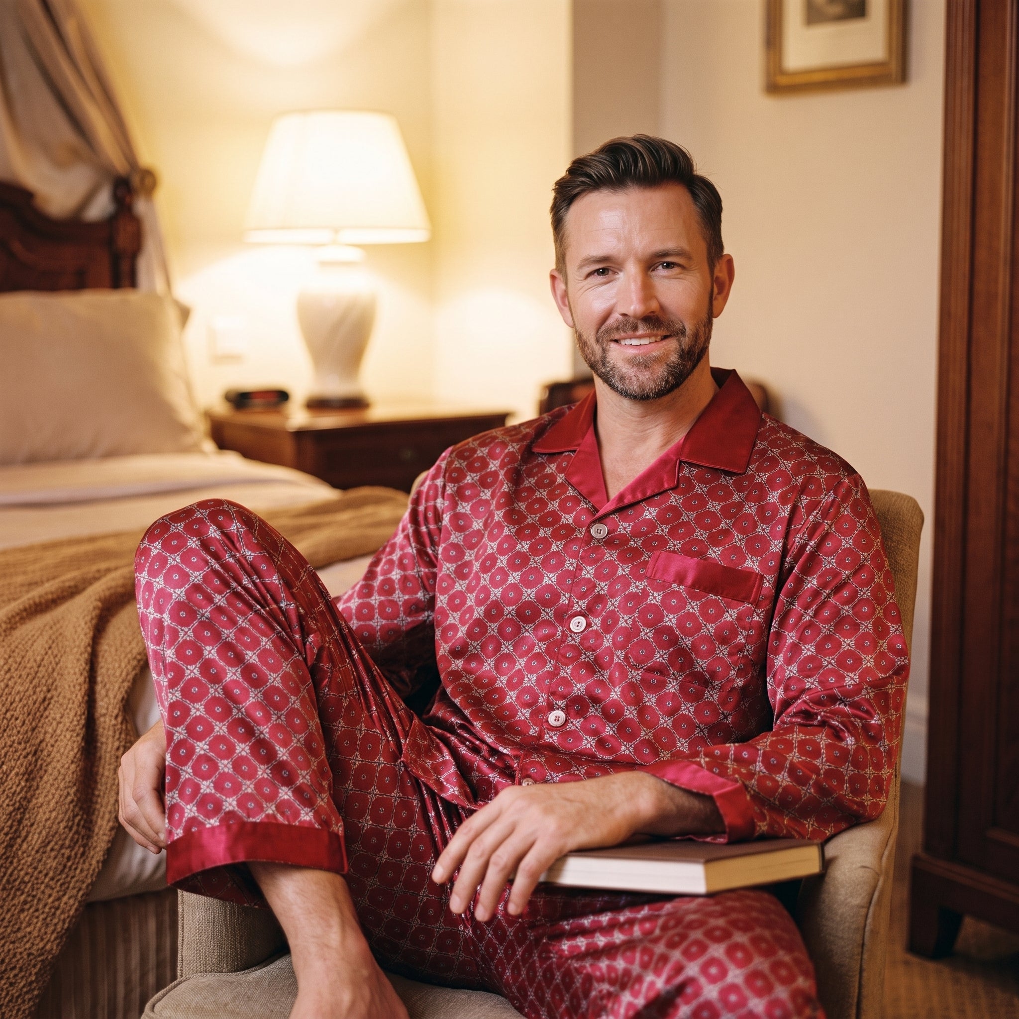 Homme installé dans un fauteuil le soir en pyjama satiné rouge, un livre ouvert sur les genoux et la lampe de chevet allumée Image