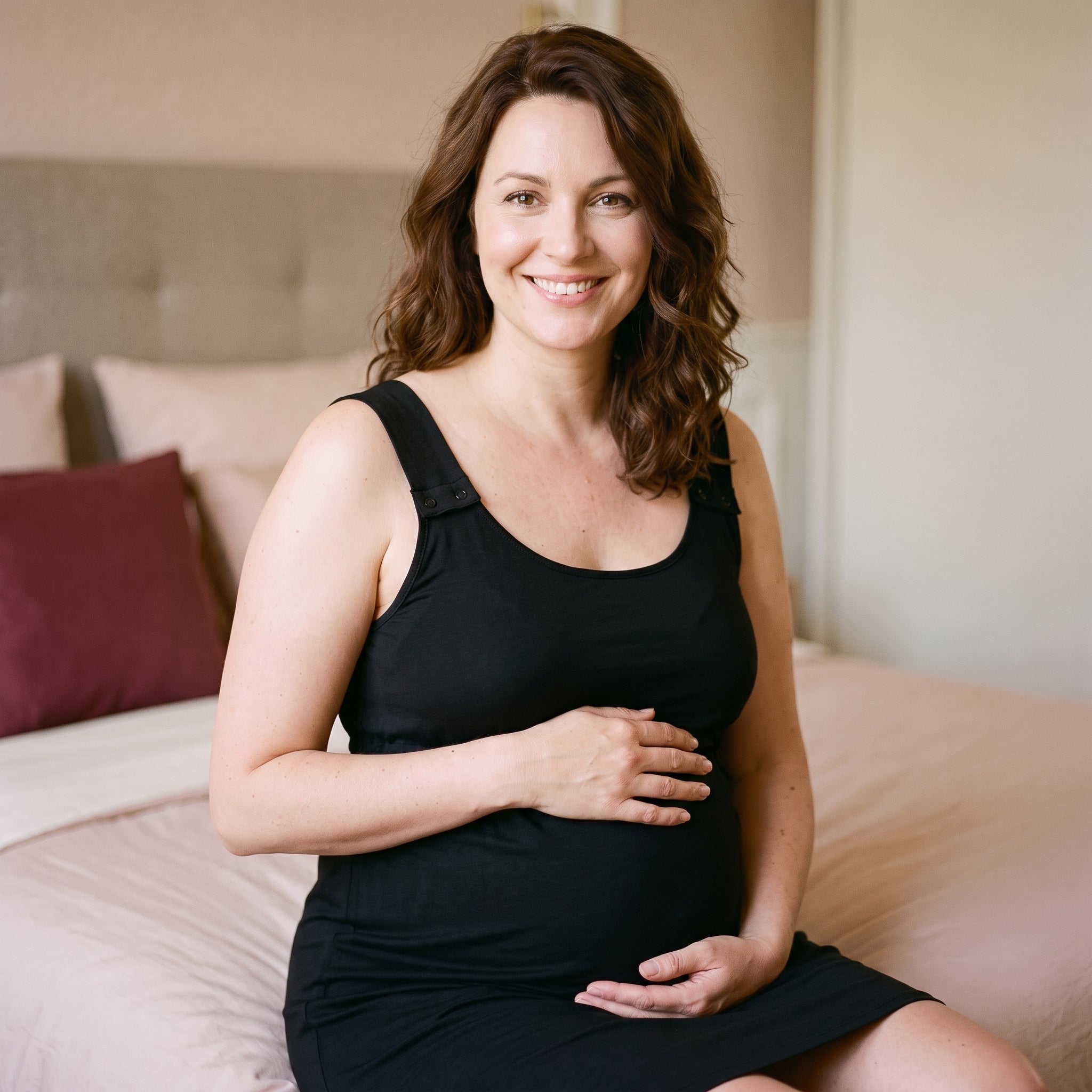Femme enceinte souriante assise sur son lit, vêtue de la nuisette de maternité noire à bretelles boutonnées Image