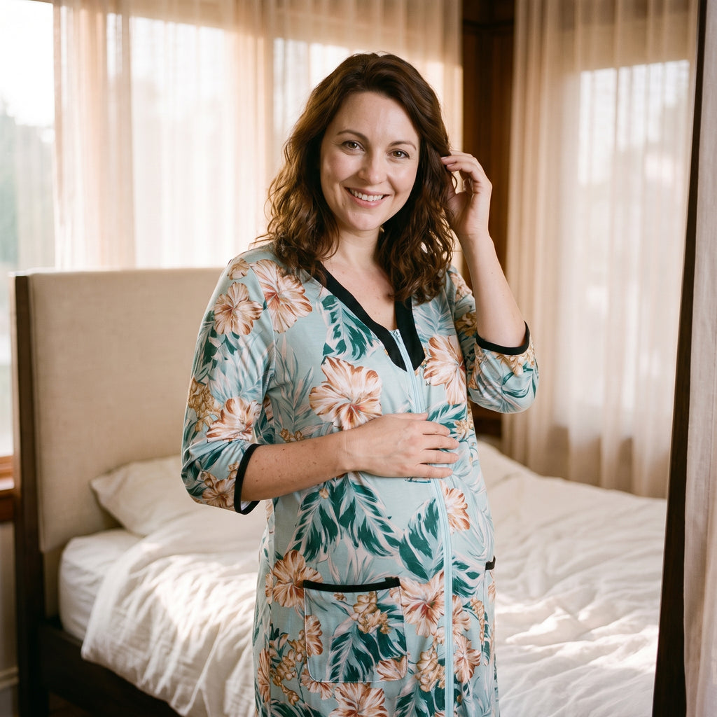 Femme enceinte debout près de la fenêtre en peignoir de maternité à fleurs, encolure en V bordée de noir et glissière remontée jusqu’à la pointe