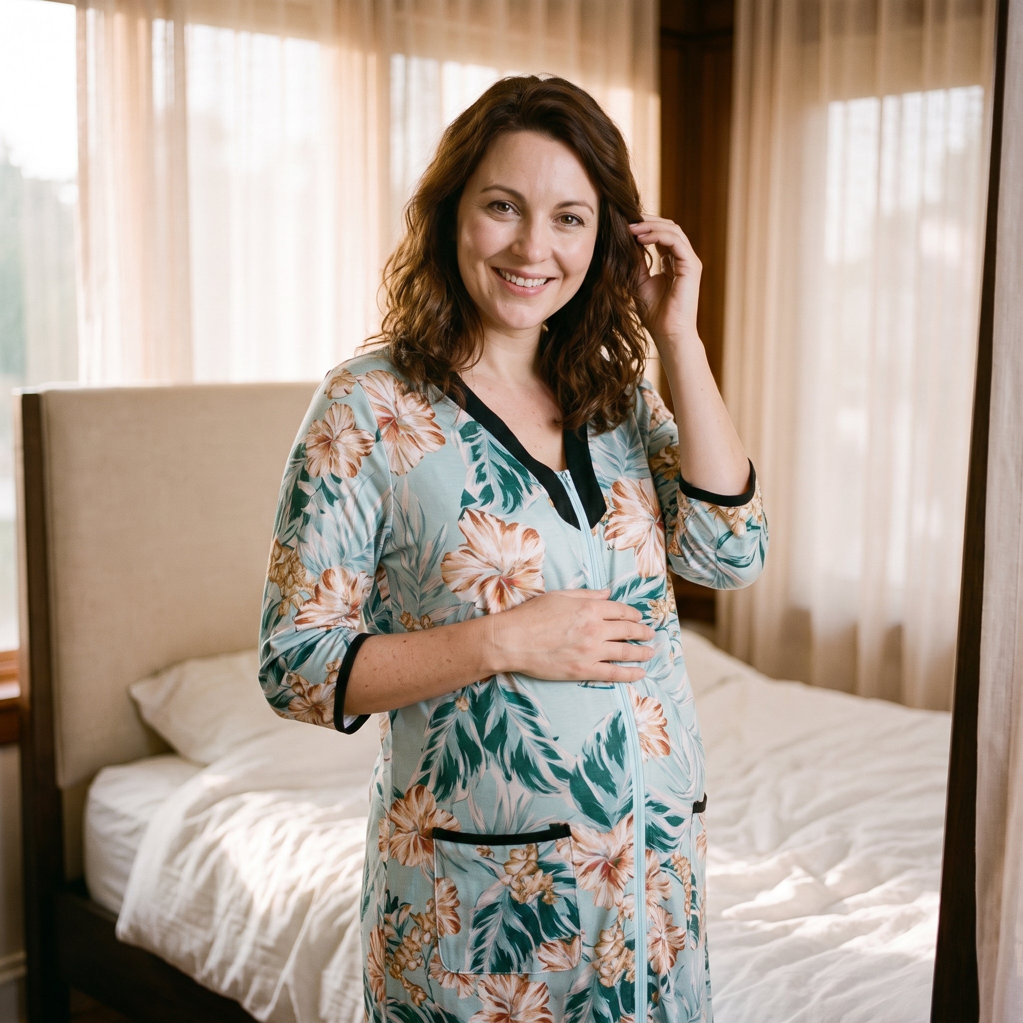 Femme enceinte debout près de la fenêtre en peignoir de maternité à fleurs, encolure en V bordée de noir et glissière remontée jusqu’à la pointe Image