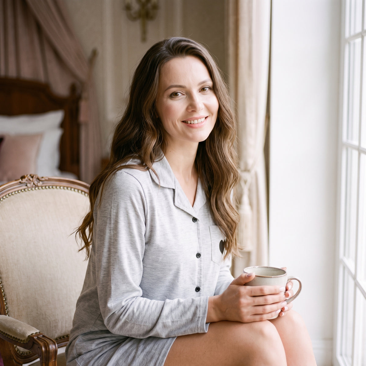 Femme souriante près de la fenêtre le matin, une tasse tenue à deux mains, en robe de chambre grise boutonnée à manches longues