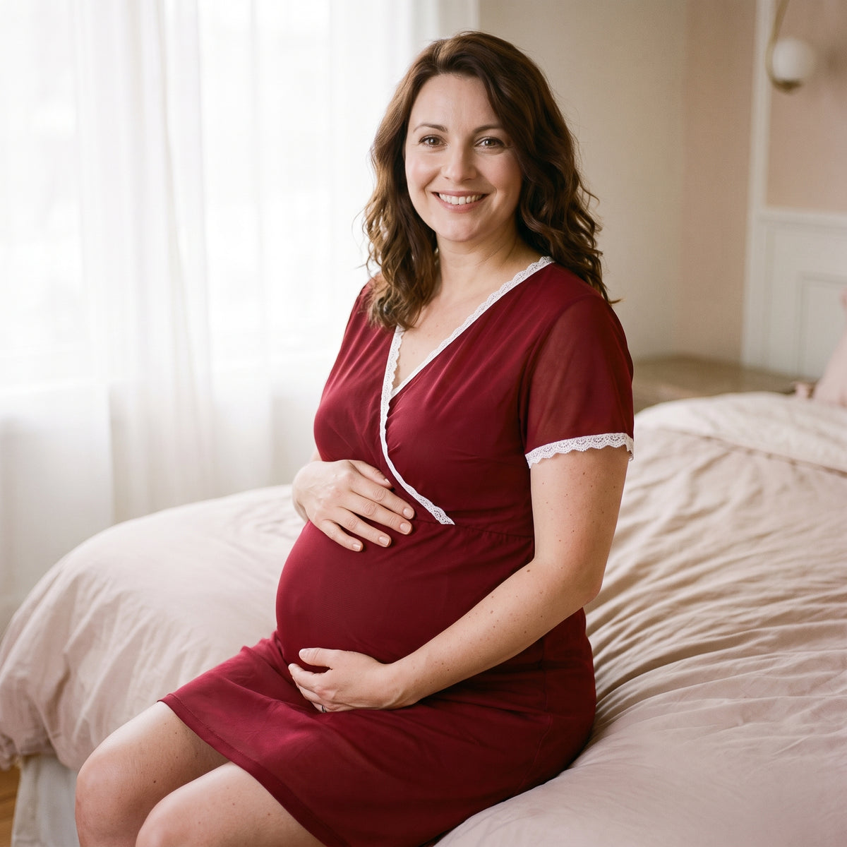Femme enceinte assise au bord du lit en nuisette de grossesse rouge, ventre rond et dentelle blanche