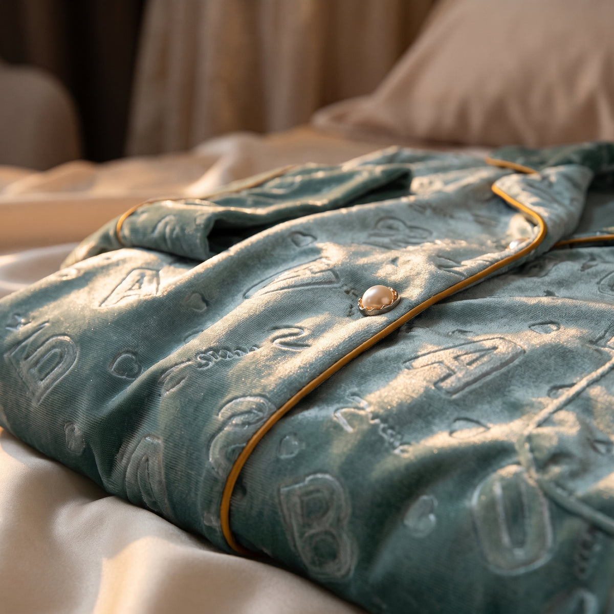 Gros plan sur le velours vert d'eau du pyjama, ses lettres en relief, son cordon doré et un bouton perle cerclé d'or