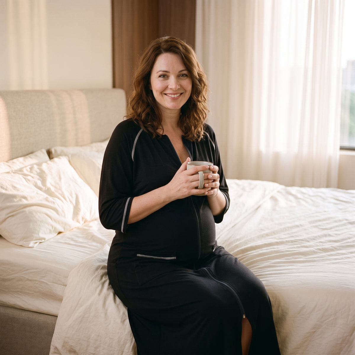 Assise au bord du lit, femme enceinte en peignoir de maternité noir, la glissière ouverte de quelques centimètres sous l’encolure ronde