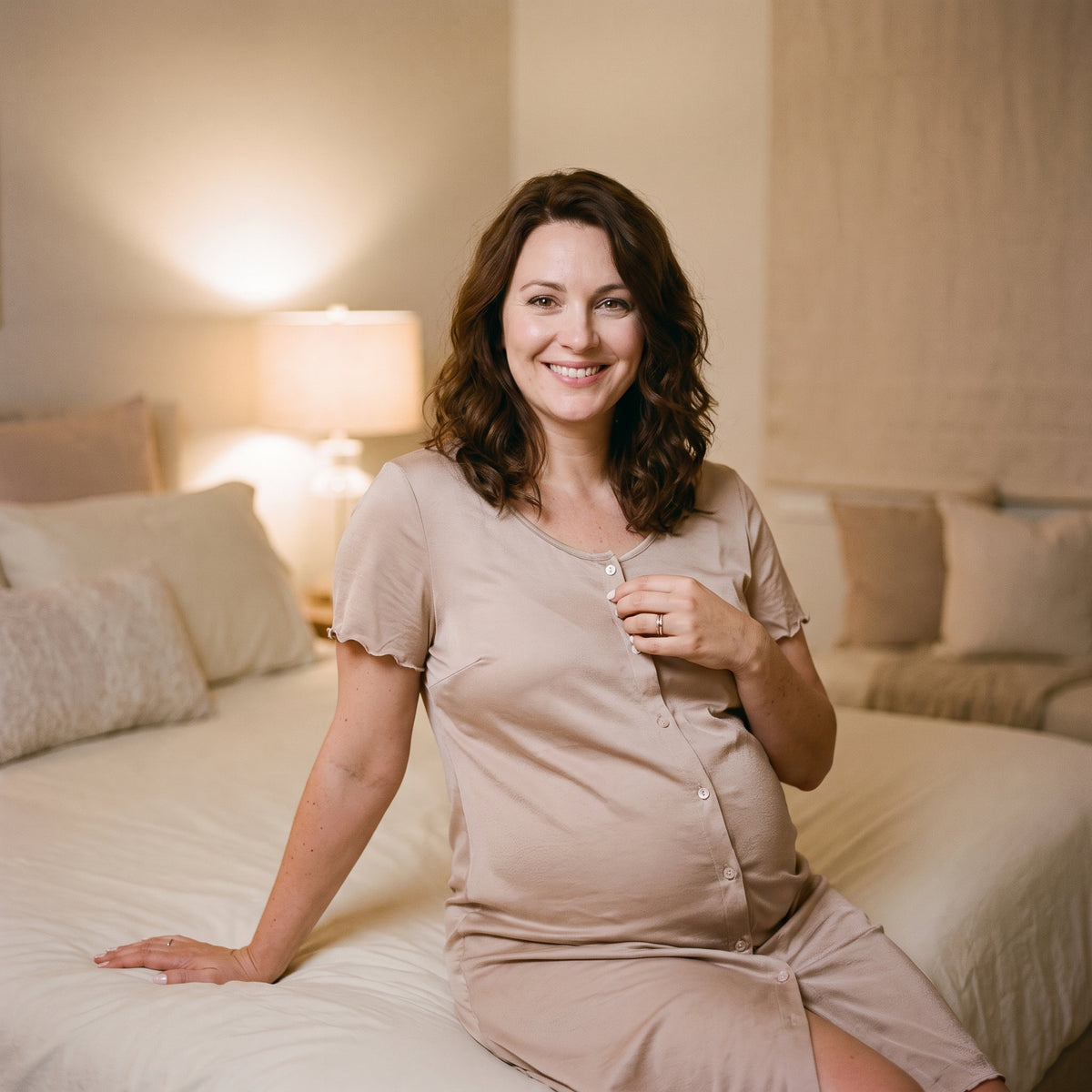 Femme enceinte assise au bord du lit le soir, la main sur un bouton du devant de sa chemise de nuit d'allaitement, lampe de chevet allumée