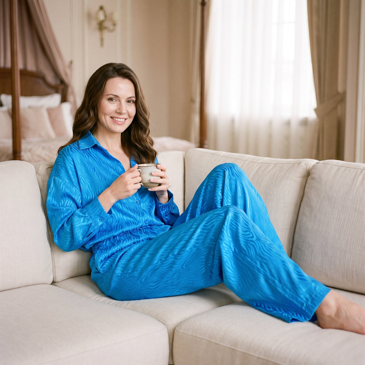Femme souriante installée sur un canapé clair, une tasse tenue à deux mains, en pyjama bleu deux pièces jusqu'à la cheville