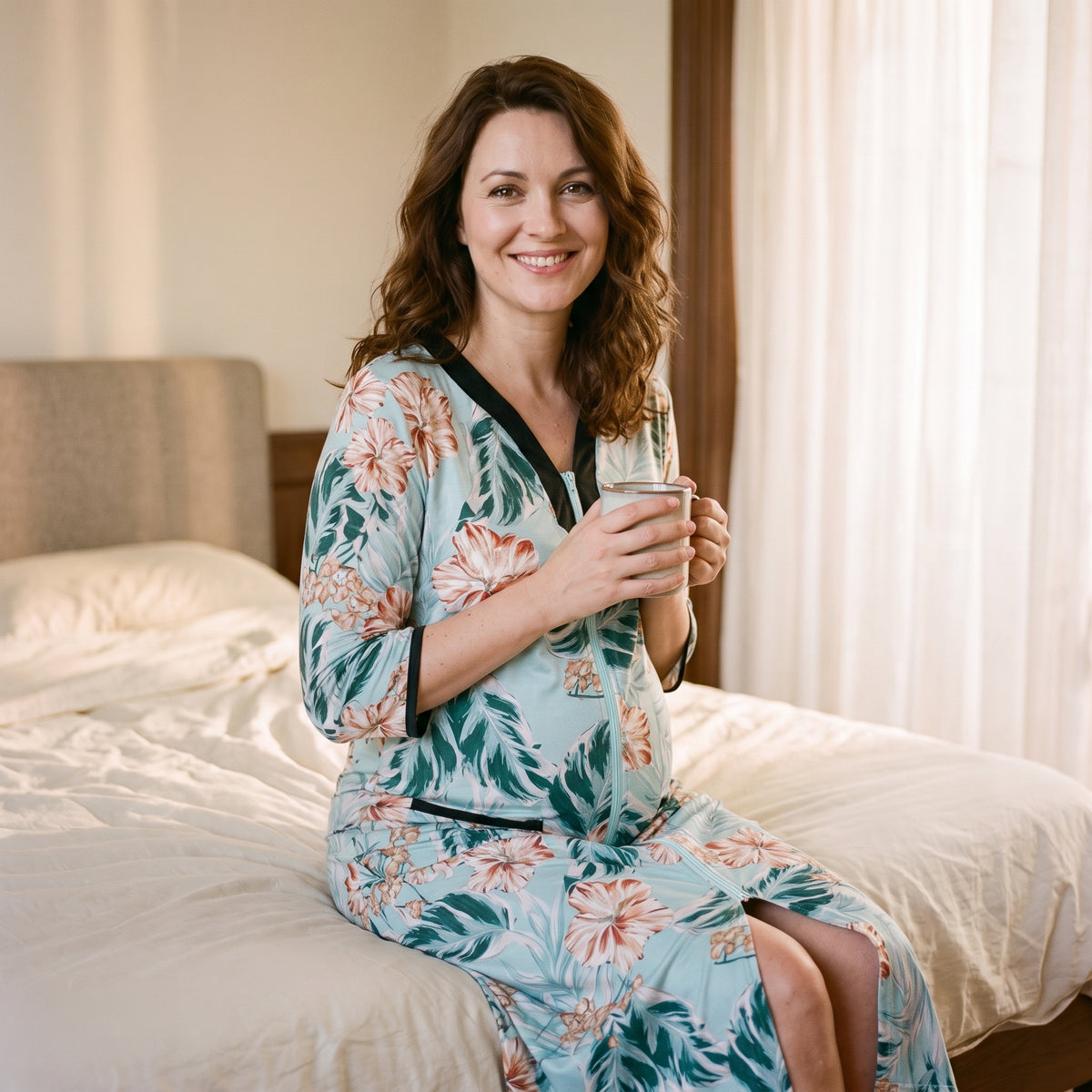 Femme enceinte assise au bord d’un lit en peignoir de maternité à fleurs, une tasse tenue à deux mains contre elle