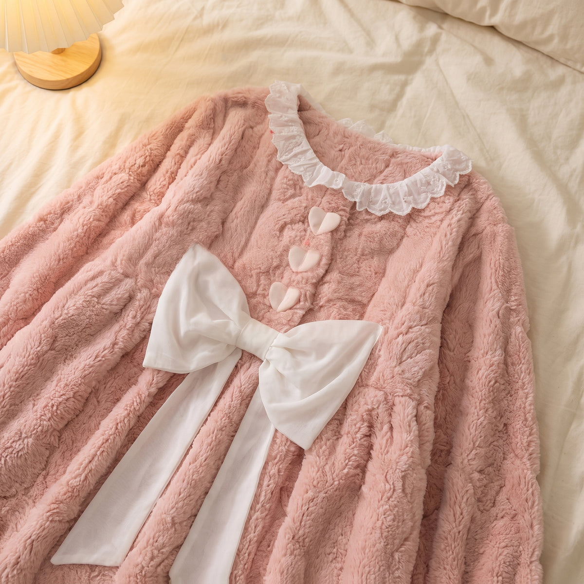 Chemise de nuit rose posée à plat sur un drap : volant de dentelle du col, trois cœurs crème alignés et grand nœud de ruban clair aux pans retombants