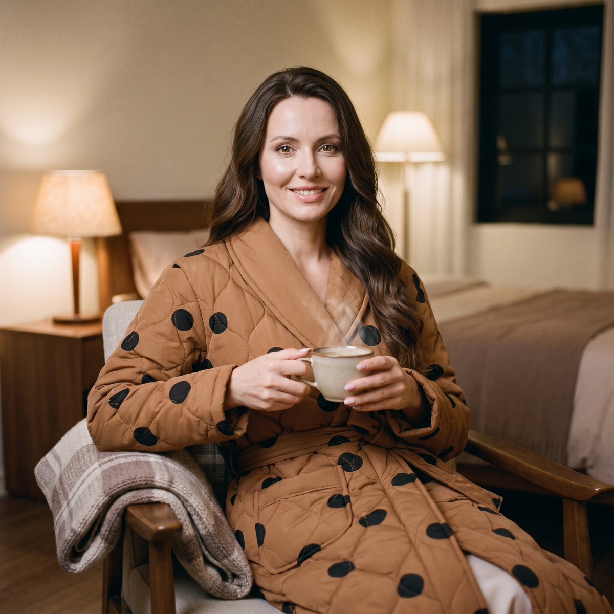 Femme assise dans un fauteuil, une tasse de café tenue à deux mains, en robe de chambre matelassée caramel à pois noirs, un plaid posé sur l’accoudoir