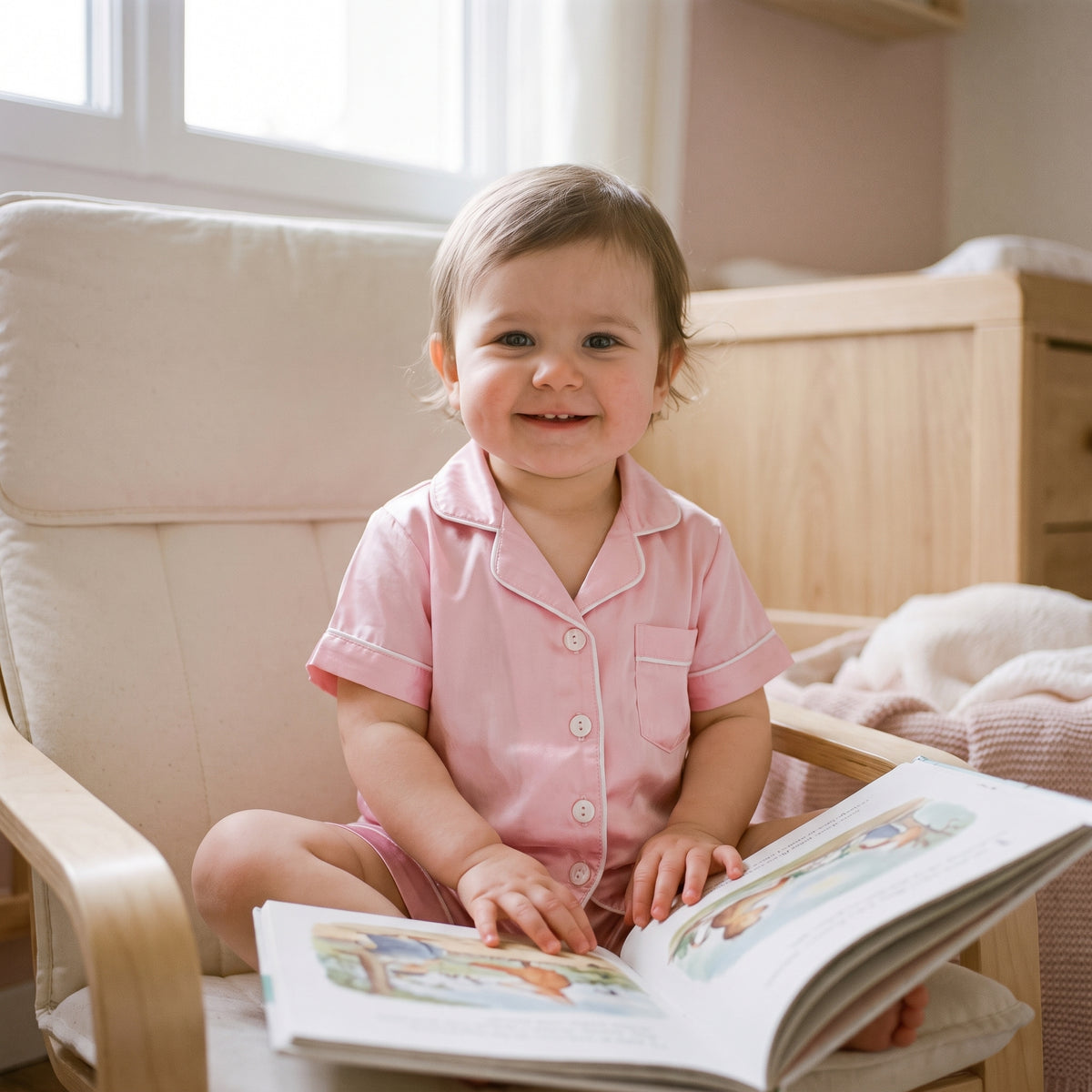 Petite fille assise dans un fauteuil clair un matin de week-end, un livre d'images ouvert sur ses genoux, en pyjama rose