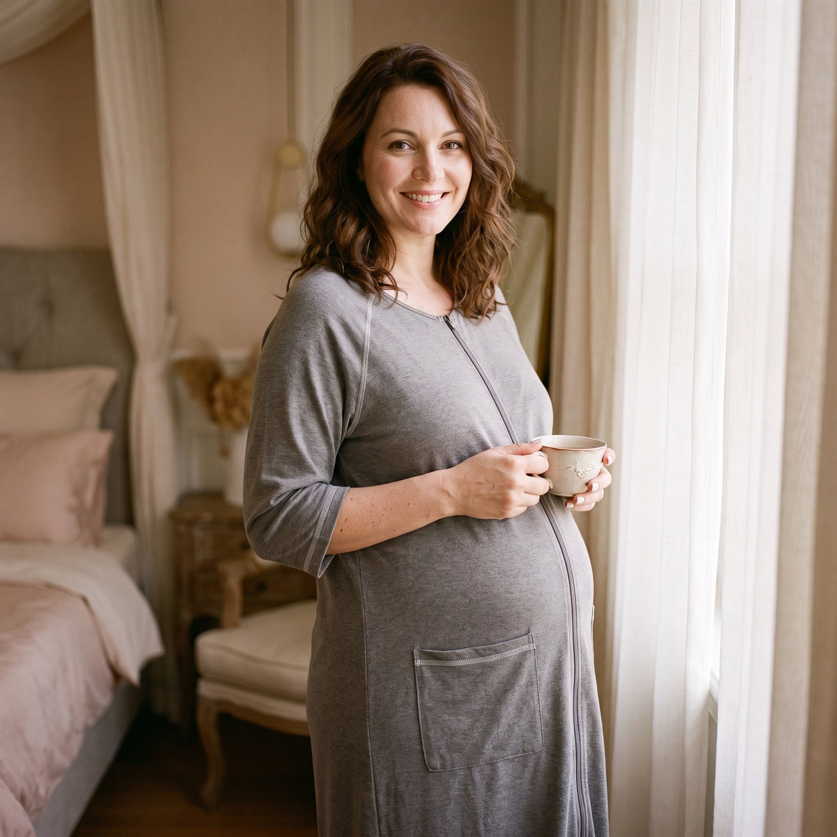 Femme debout près d'une fenêtre voilée, une tasse tenue à deux mains, en robe de chambre grise zippée