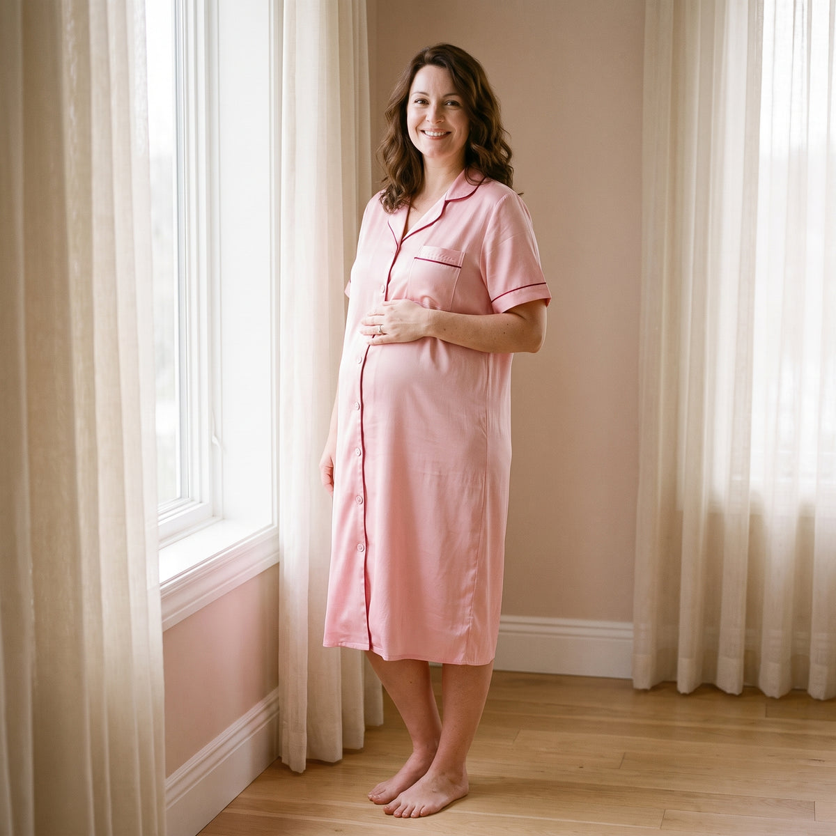Chemise de nuit de maternité rose poudrée portée debout, ourlet à mi-mollet, devant une fenêtre au petit matin