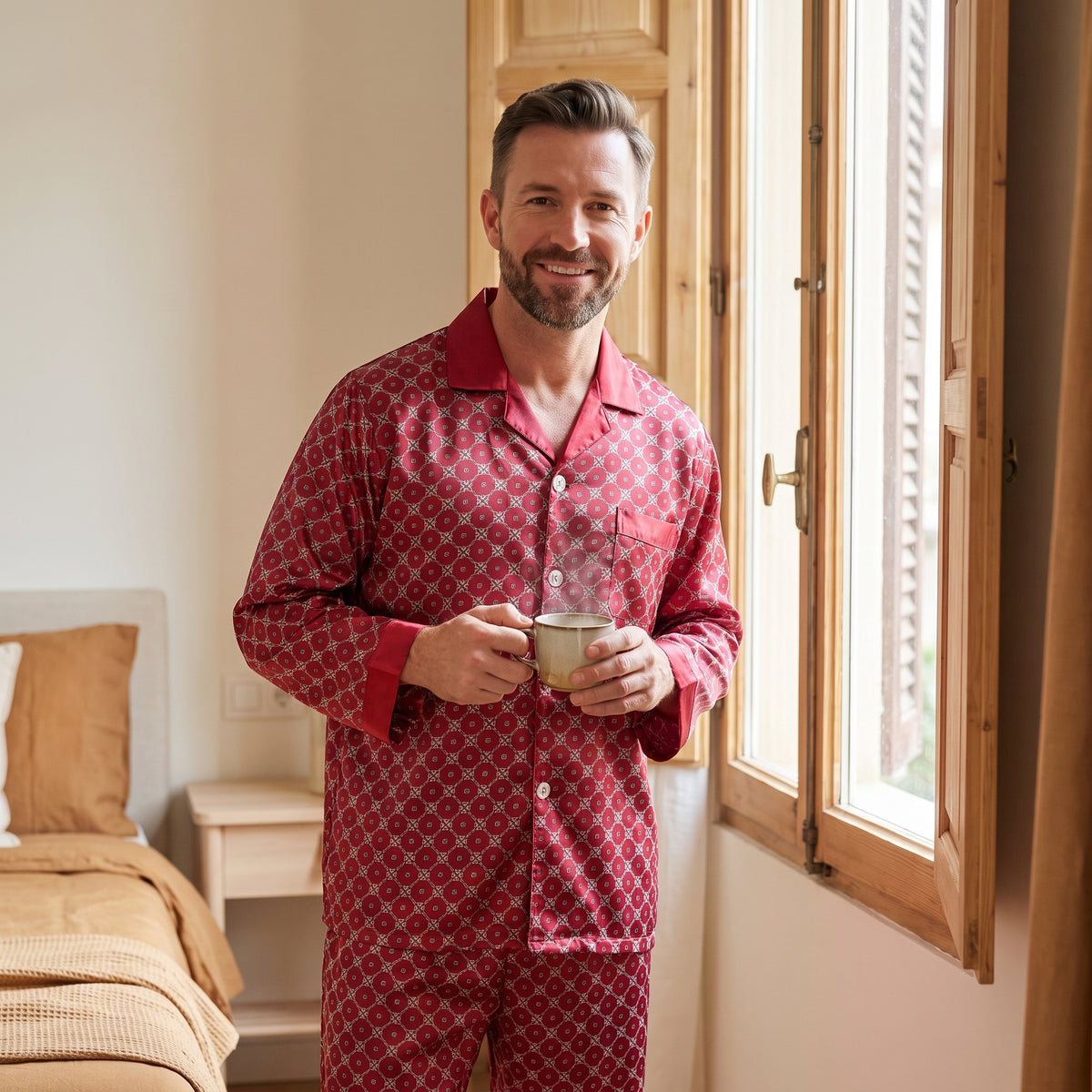 Homme devant la fenêtre aux volets ouverts le matin, une tasse de café entre les mains, en pyjama satiné rouge à motifs
