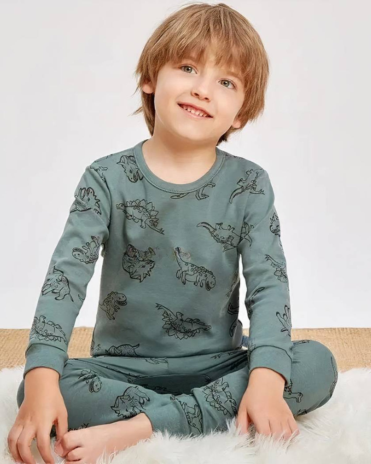 Garcon souriant en pyjama dinosaures vert sauge, assis en tailleur