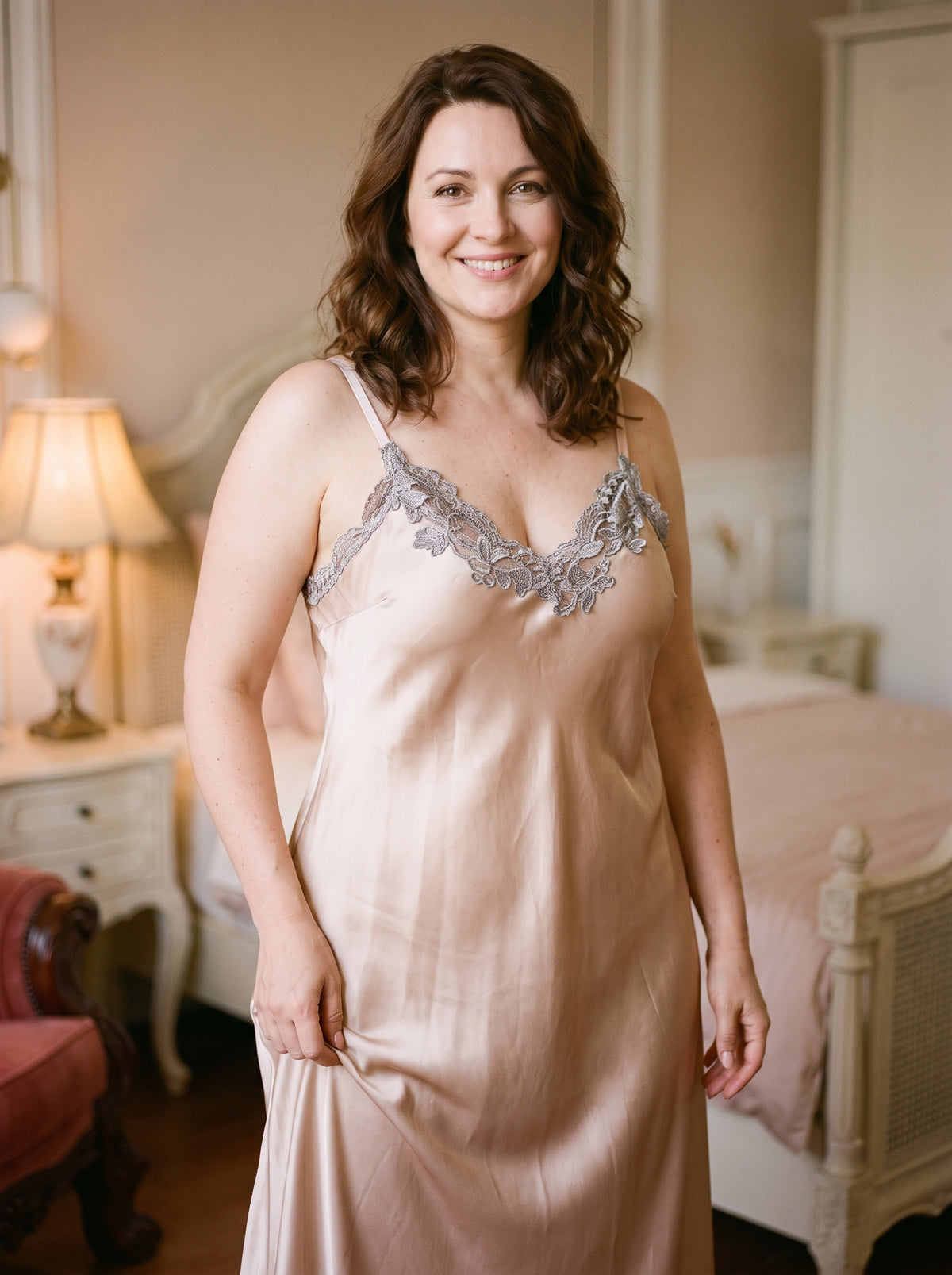 Femme souriante en nuisette longue de satin beige champagne, fines bretelles posées sur les épaules, décolleté en V bordé d’une guipure argentée
