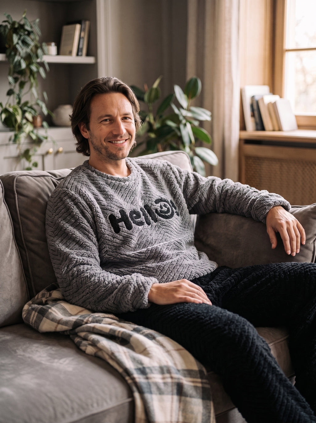 Homme portant le pyjama d'hiver en peluche, sweat gris à lettrage Hello et pantalon noir, installé sur un canapé