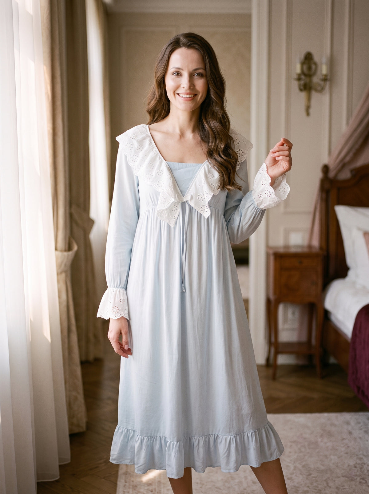 Femme souriante en nuisette longue bleu pâle, le grand col de broderie anglaise posé sur les épaules et le volant de l’ourlet au mollet