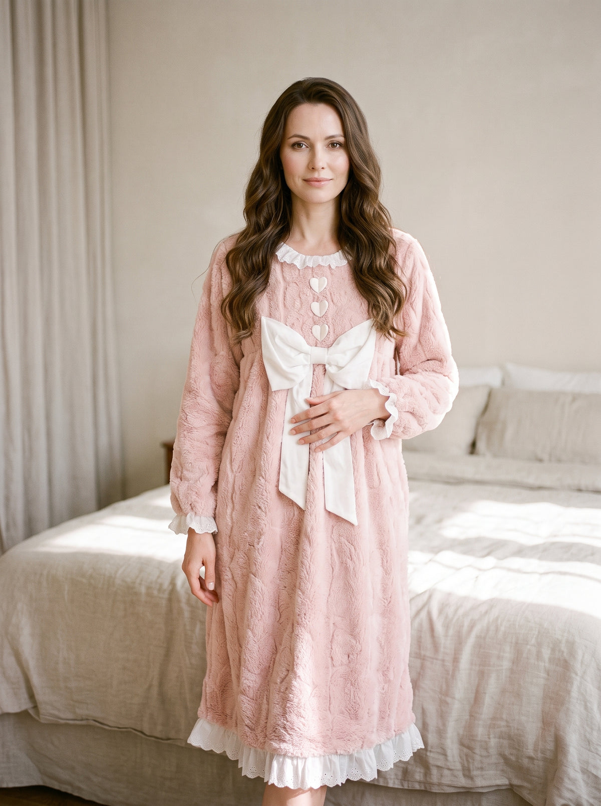 Femme souriante debout dans une chambre en chemise de nuit rose duveteuse, col et poignets à volant de dentelle blanche, trois cœurs et grand nœud sur le devant, ourlet au mollet