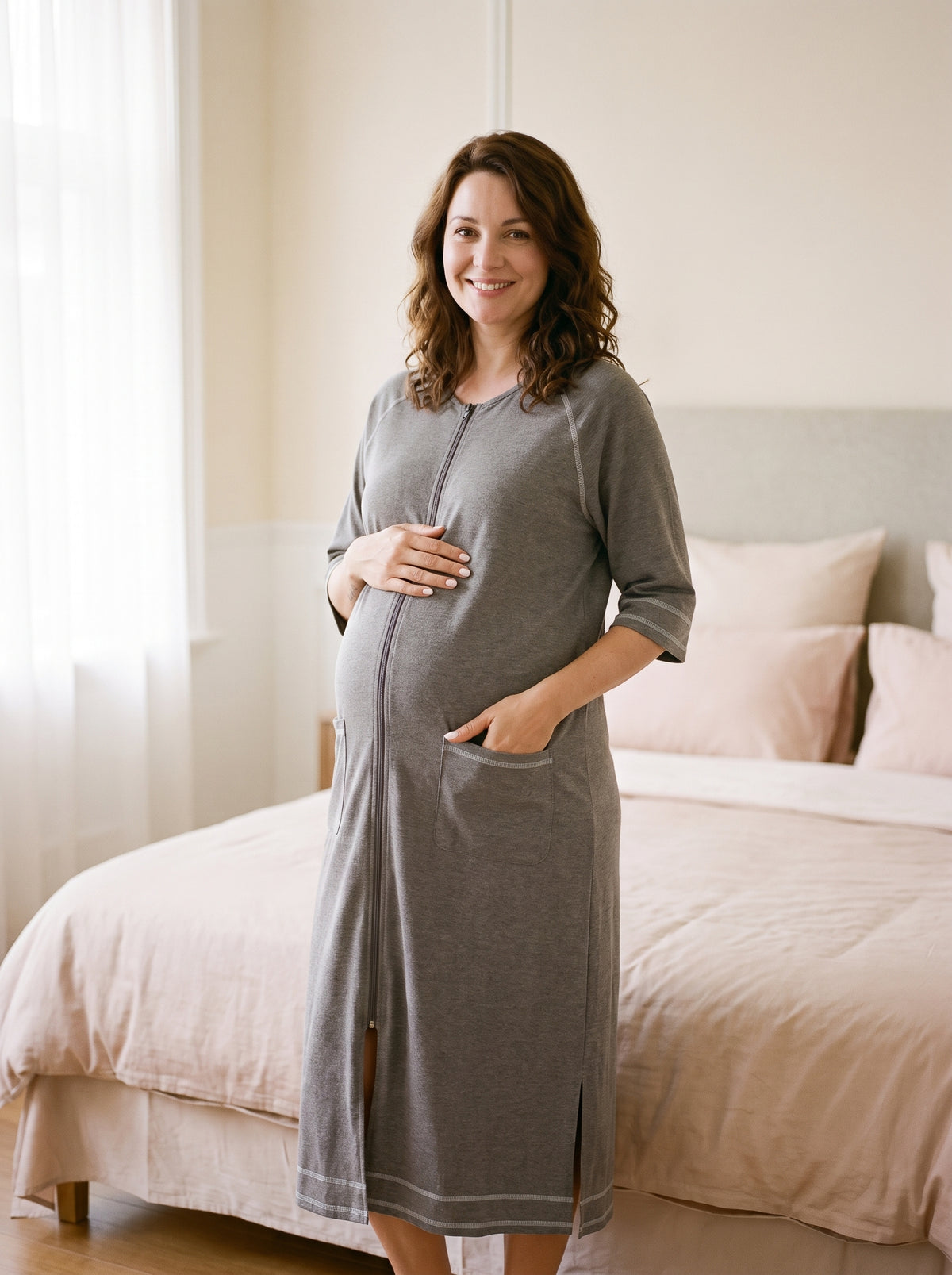 Future maman souriante debout près d'un lit en robe de chambre grise zippée, une main posée sur son ventre rond