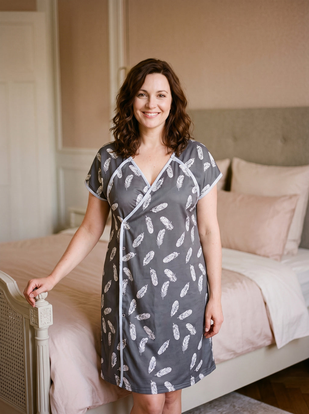 Chemise de nuit d'allaitement grise à imprimé de plumes portée de face au pied d'un lit, pan croisé à boutons-pression et manches courtes