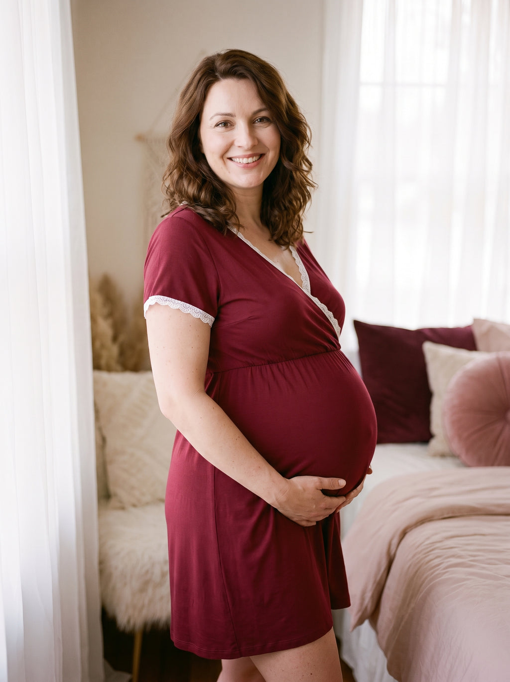 Future maman souriante en nuisette de grossesse rouge à décolleté croisé, debout près du lit