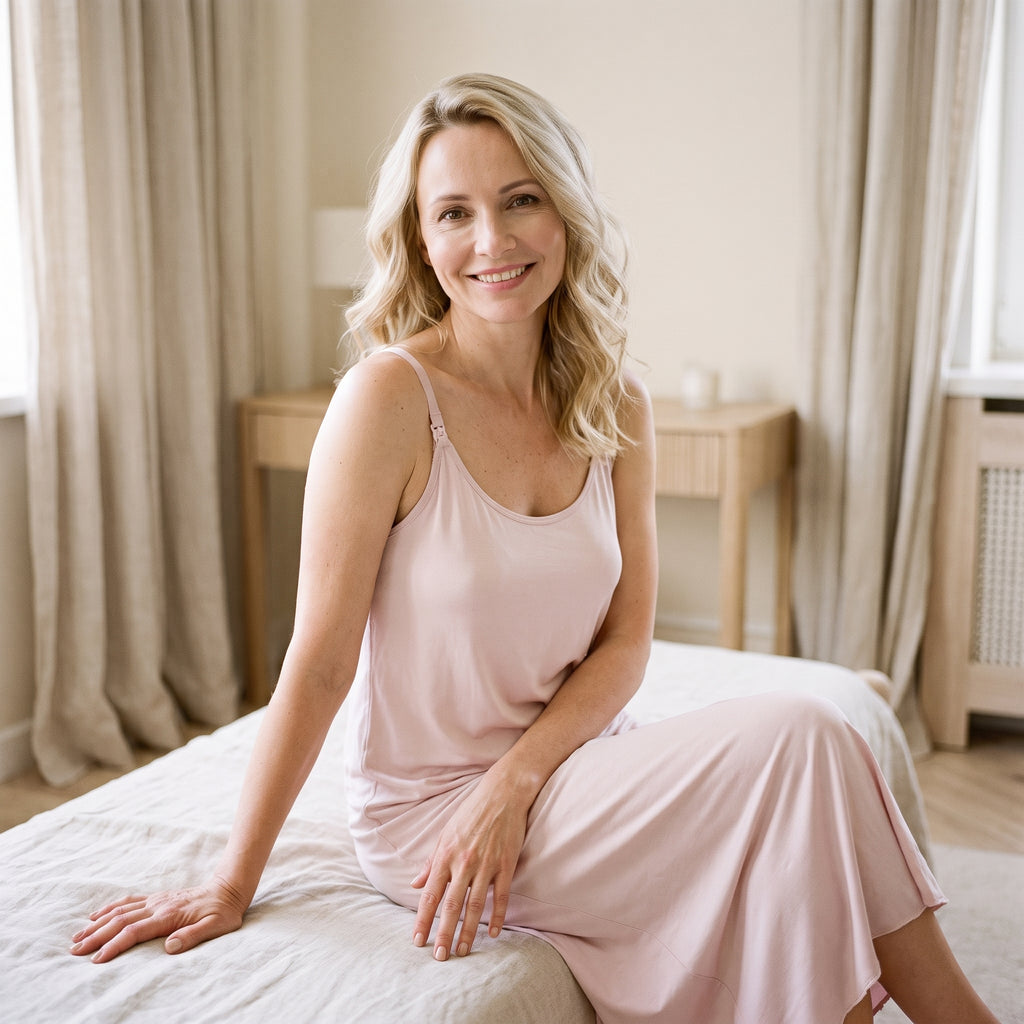 Femme souriante assise sur son lit en nuisette d’allaitement rose, la douceur du tissu sur la peau et le tombé souple du rose poudré
