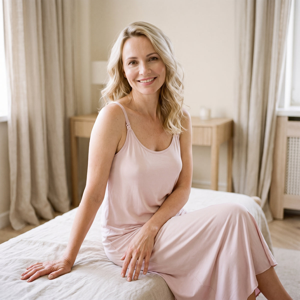 Femme souriante assise sur son lit en nuisette d’allaitement rose, la douceur du tissu sur la peau et le tombé souple du rose poudré