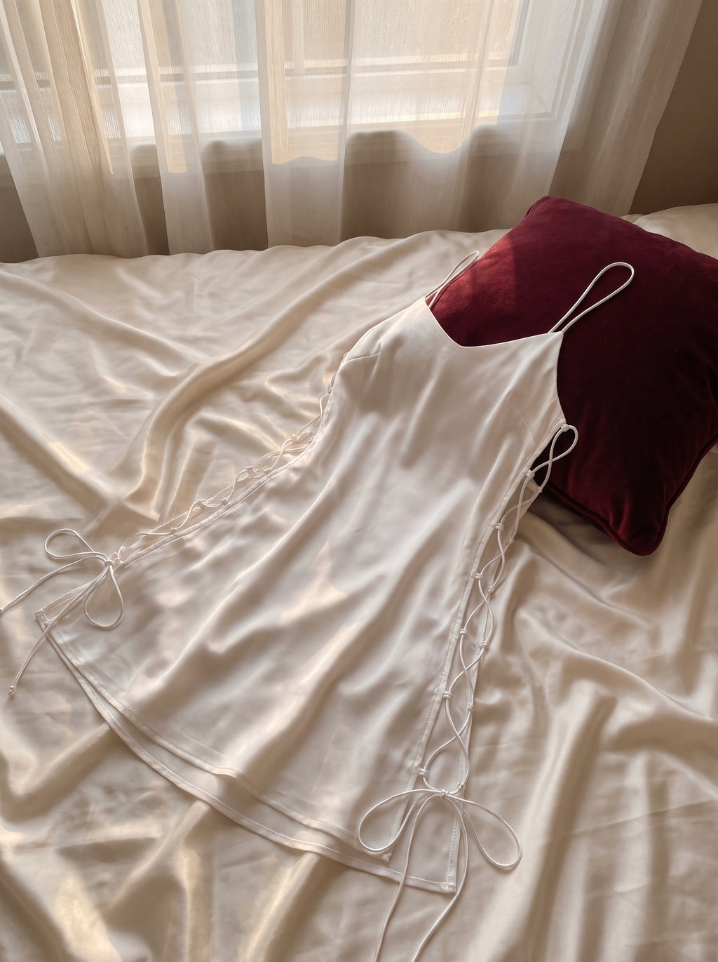 Nuisette blanche en satin deposee sur lit aux draps ivoire, coussin bordeaux, decor boudoir intime