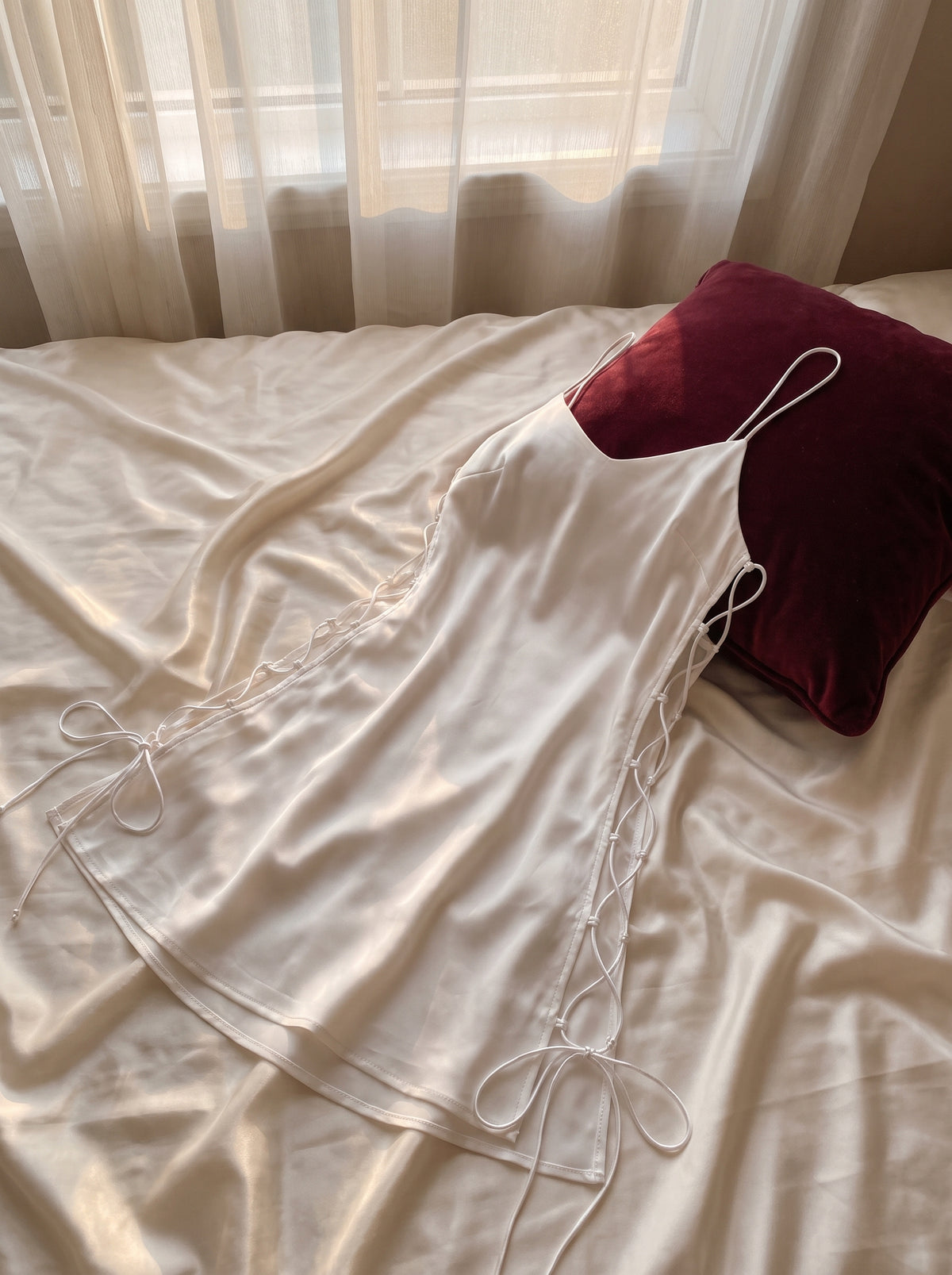Nuisette blanche en satin deposee sur lit aux draps ivoire, coussin bordeaux, decor boudoir intime