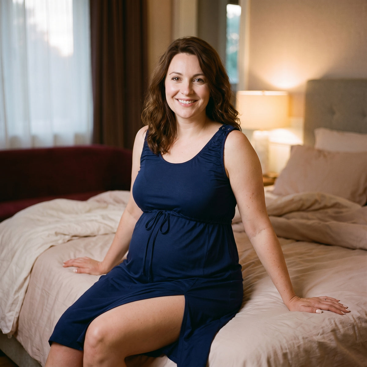 Femme enceinte assise au bord du lit en nuisette de grossesse et d'allaitement bleu nuit, fente latérale ouverte sur la cuisse, cordon noué sous la poitrine