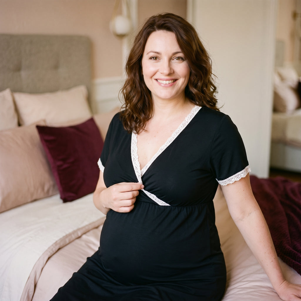 Femme enceinte assise sur le lit, la main au bord de dentelle du décolleté croisé de sa nuisette de maternité noire