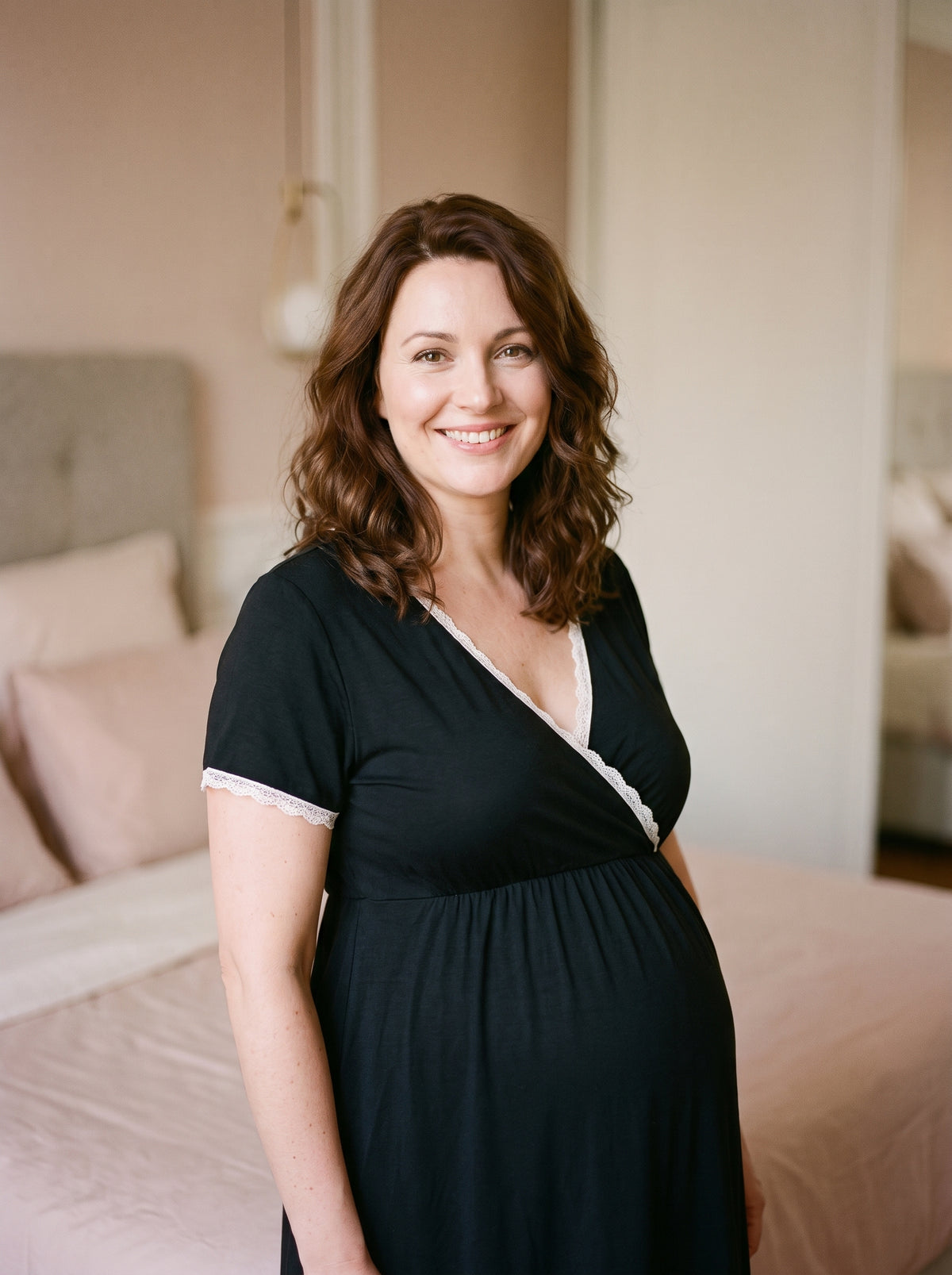 Femme enceinte souriante debout dans une chambre, en nuisette de maternité noire à dentelle blanche, le ventre rond sous la couture empire
