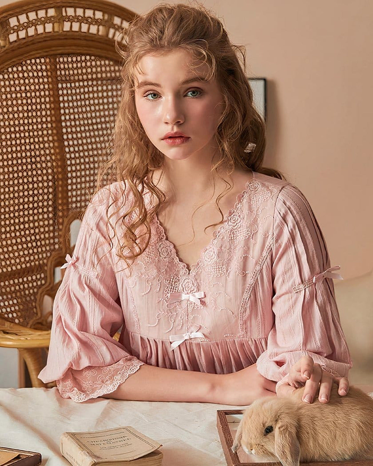 Femme assise en nuisette vintage rose, visage visible, décolleté en V et deux nœuds ivoire sur le buste