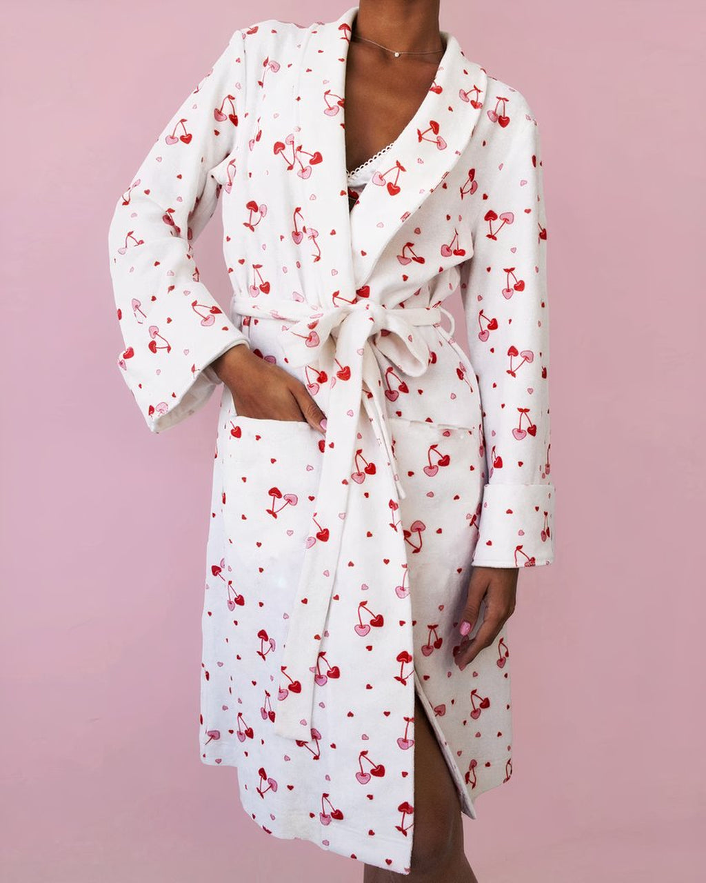 Peignoir long blanc à cerises porté devant un fond rose, la ceinture nouée à la taille et une main glissée dans la poche plaquée