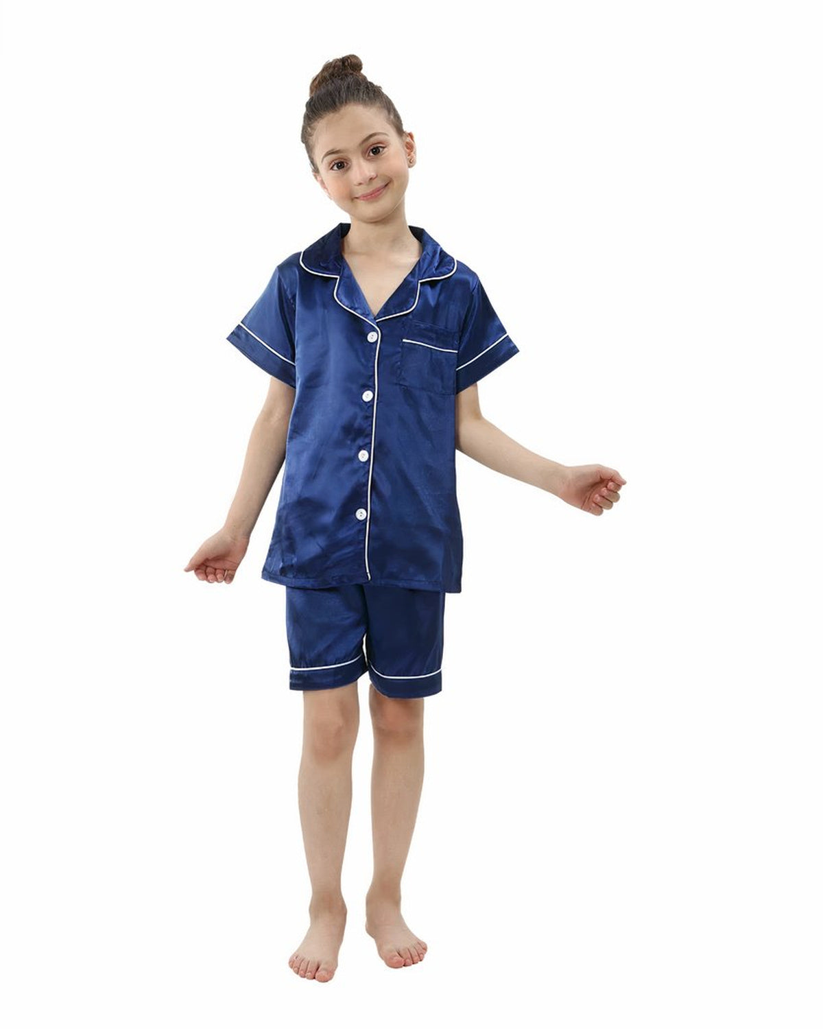Petite fille souriante debout en pyjama deux pièces bleu marine satiné, chemise à manches courtes boutonnée et short assorti bordés d'un liseré blanc