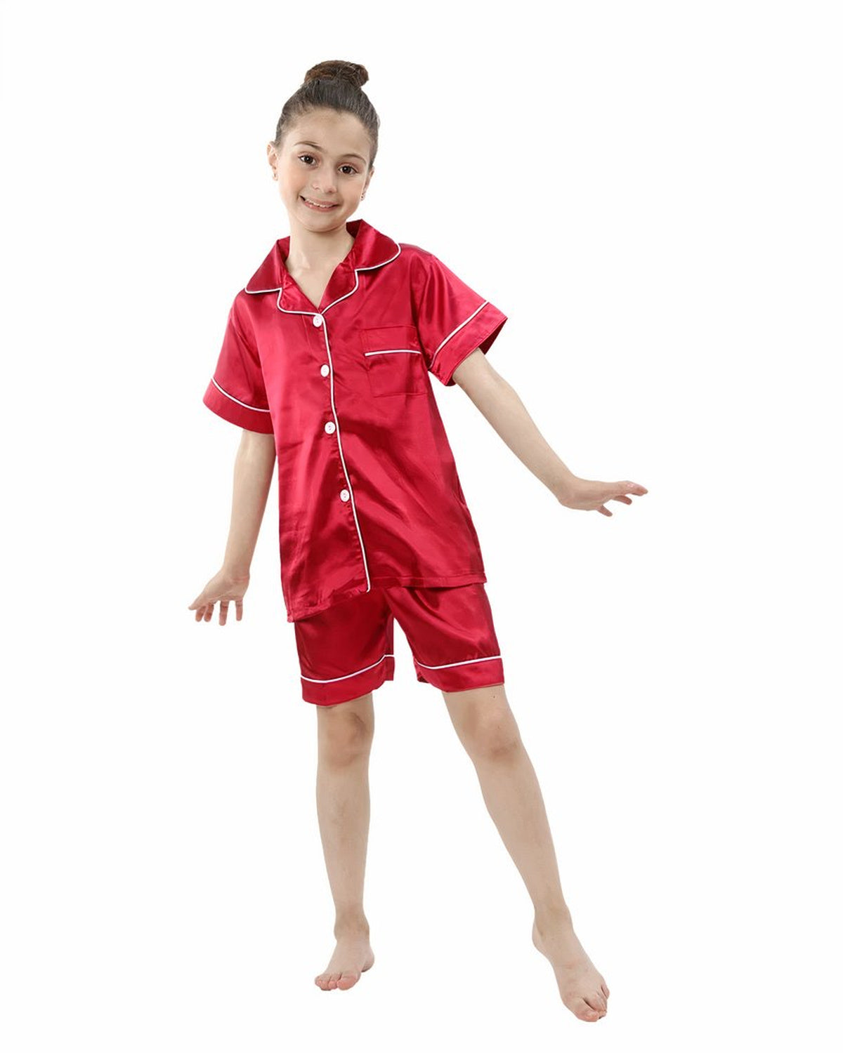 Petite fille souriante debout en pyjama deux pièces rouge satiné, chemise à manches courtes boutonnée et short assorti bordés d'un liseré blanc
