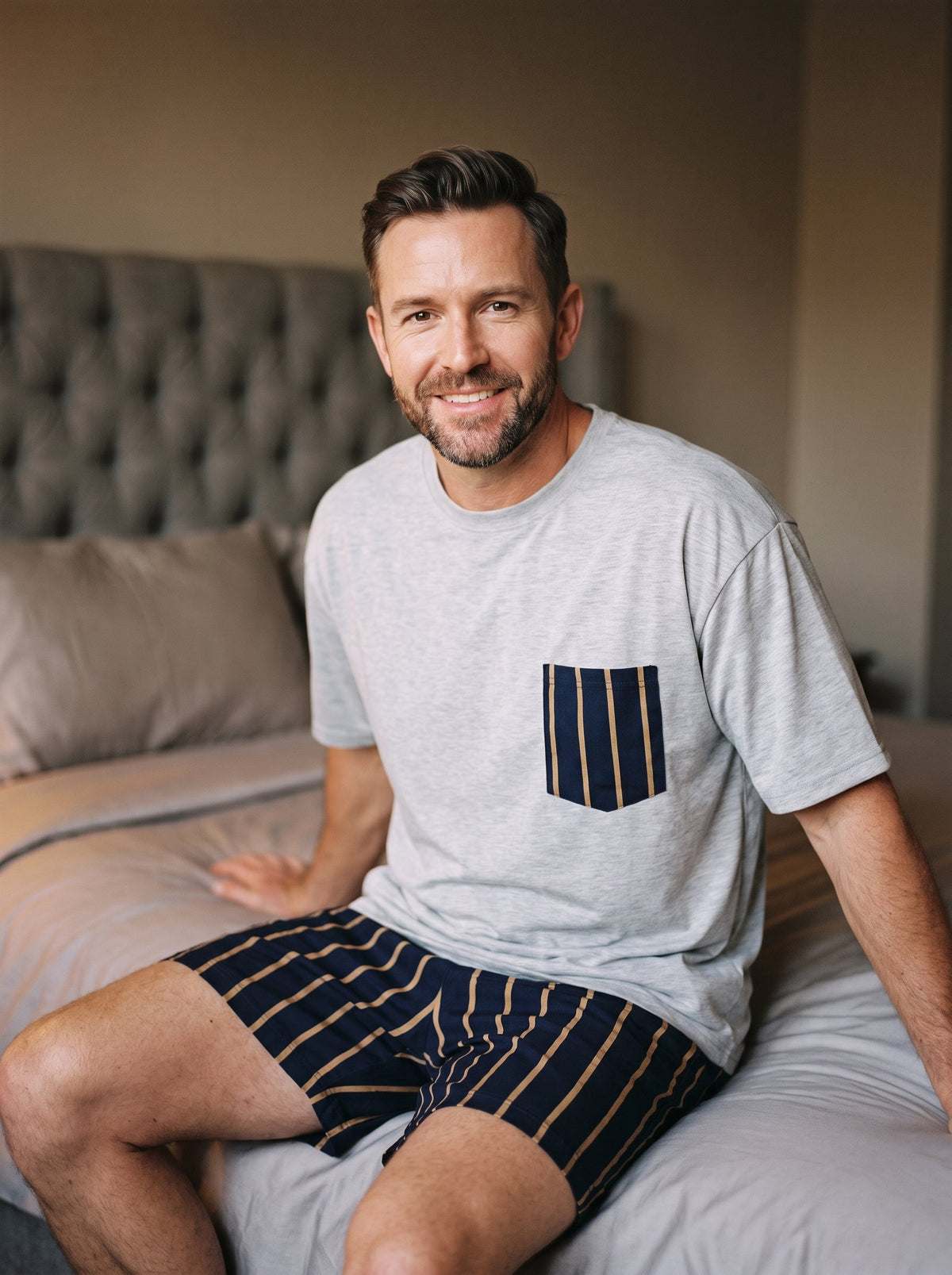 Homme souriant portant le pyjama été gris chiné, poche rayée bleu marine et short coordonné, assis sur un lit