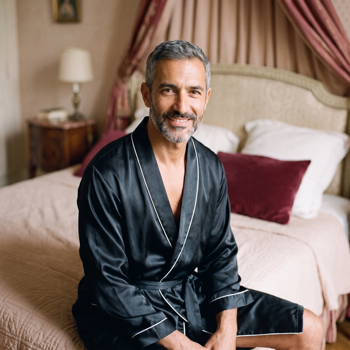 Homme souriant assis au bord du lit avec une robe de chambre noire satinée