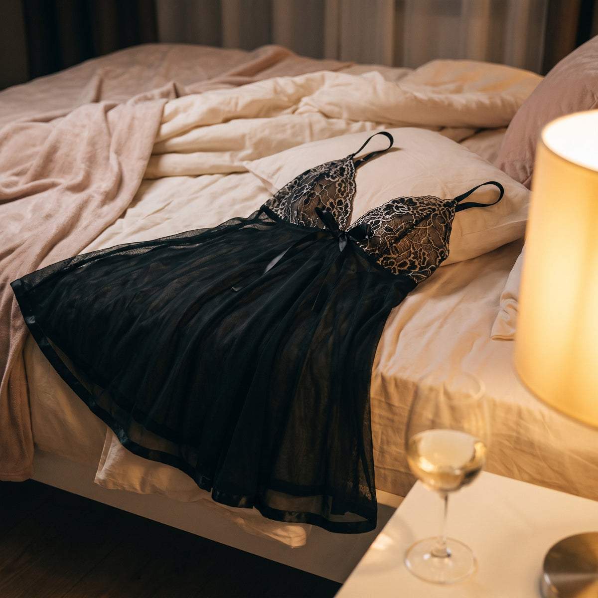 Nuisette noire posée sur un lit défait, lampe de chevet allumée et verre à pied posé sur la table de nuit