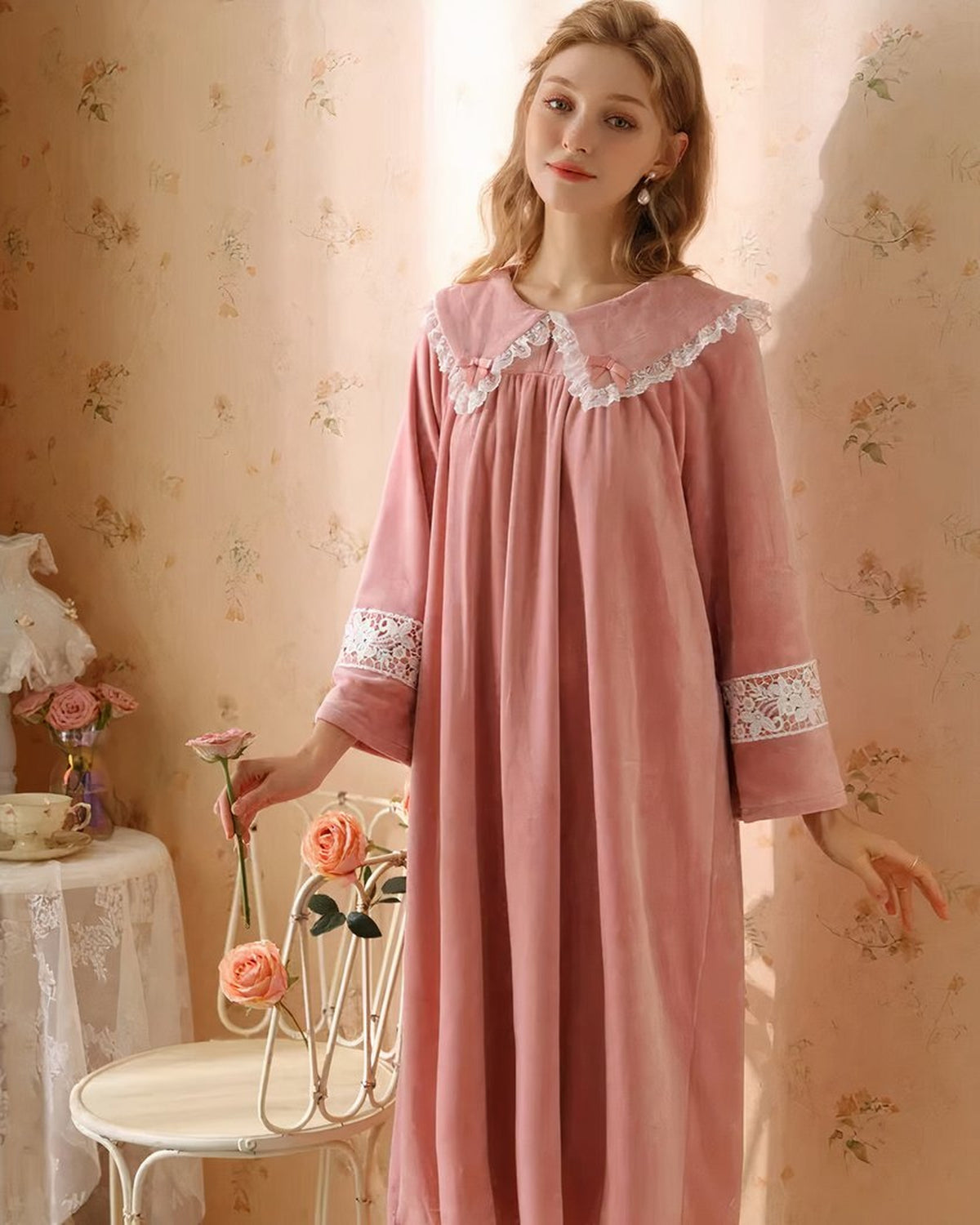 Modele souriante portant la robe de nuit princesse en velours rose, debout de face tenant une rose, decor romantique