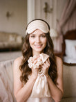 Femme brune souriante avec le kit de nuit en soie champagne, masque relevé sur le front, ambiance boudoir