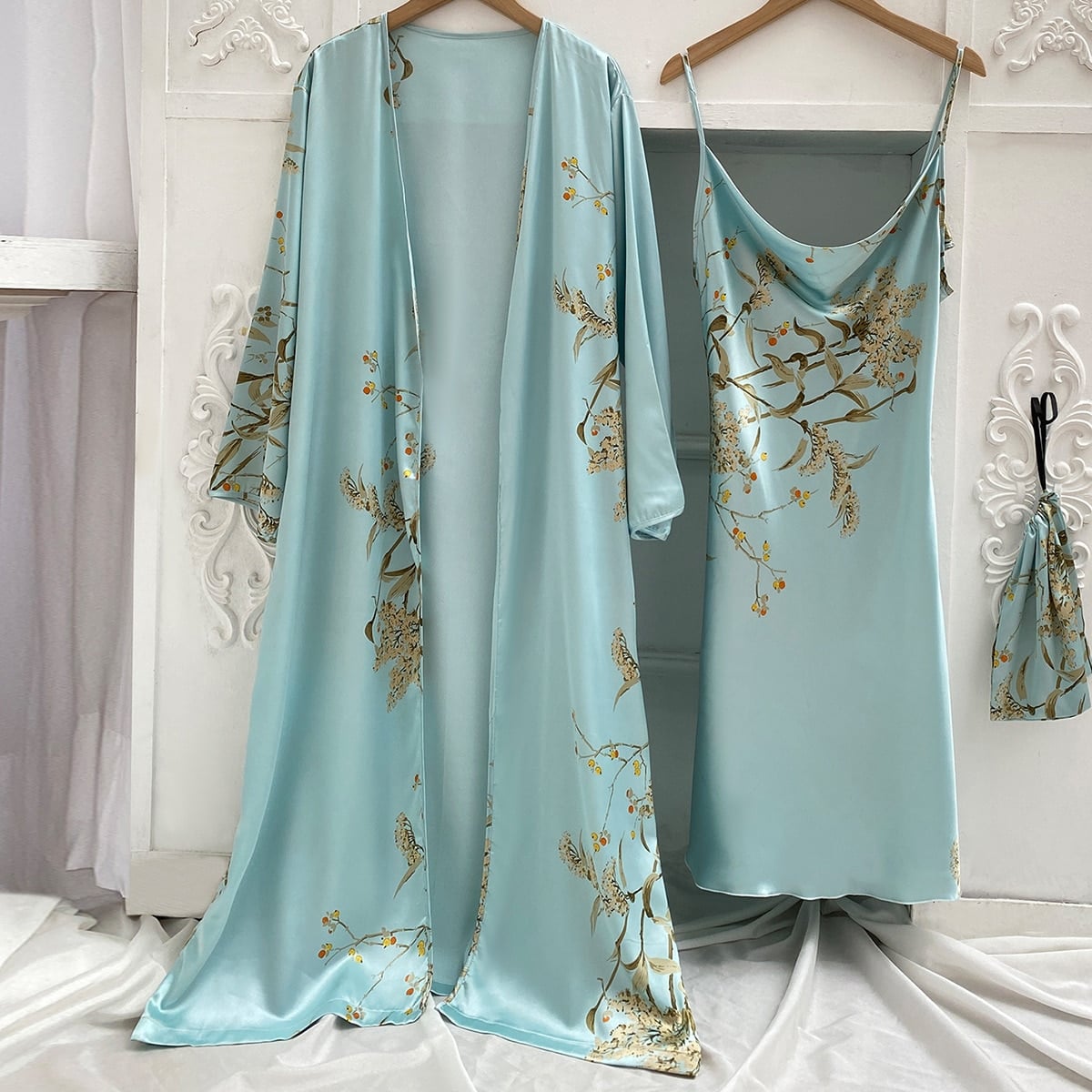 Un kimono et une nuisette en satin bleu clair avec des motifs floraux accrochés sur des cintres côte à côte sur un mur blanc avec des moulures. A côté de la nuisette, il y a un petit sac du même motif accroché à une patère.
