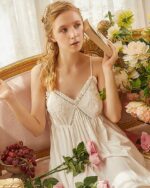 Femme blonde portant une nuisette blanche bohème, assise sur un canapé, avec des fleurs et un livre