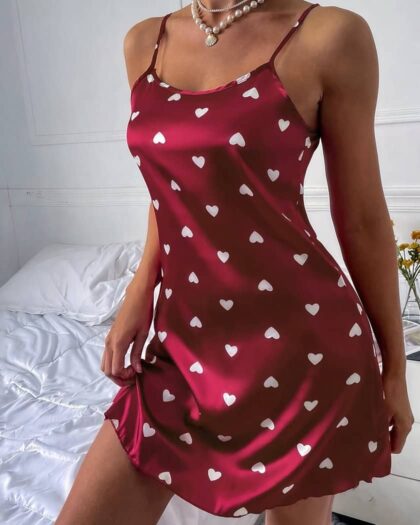 Corps de femme debout devant un lit avec draps blancs portant une nuisette courte en satin rouge avec un imprimé de petits cœurs blanc.
