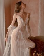 Femme blonde portant une nuisette transparente blanche près d'un canapé rouge avec des rideaux blancs transparents aux fenêtres.